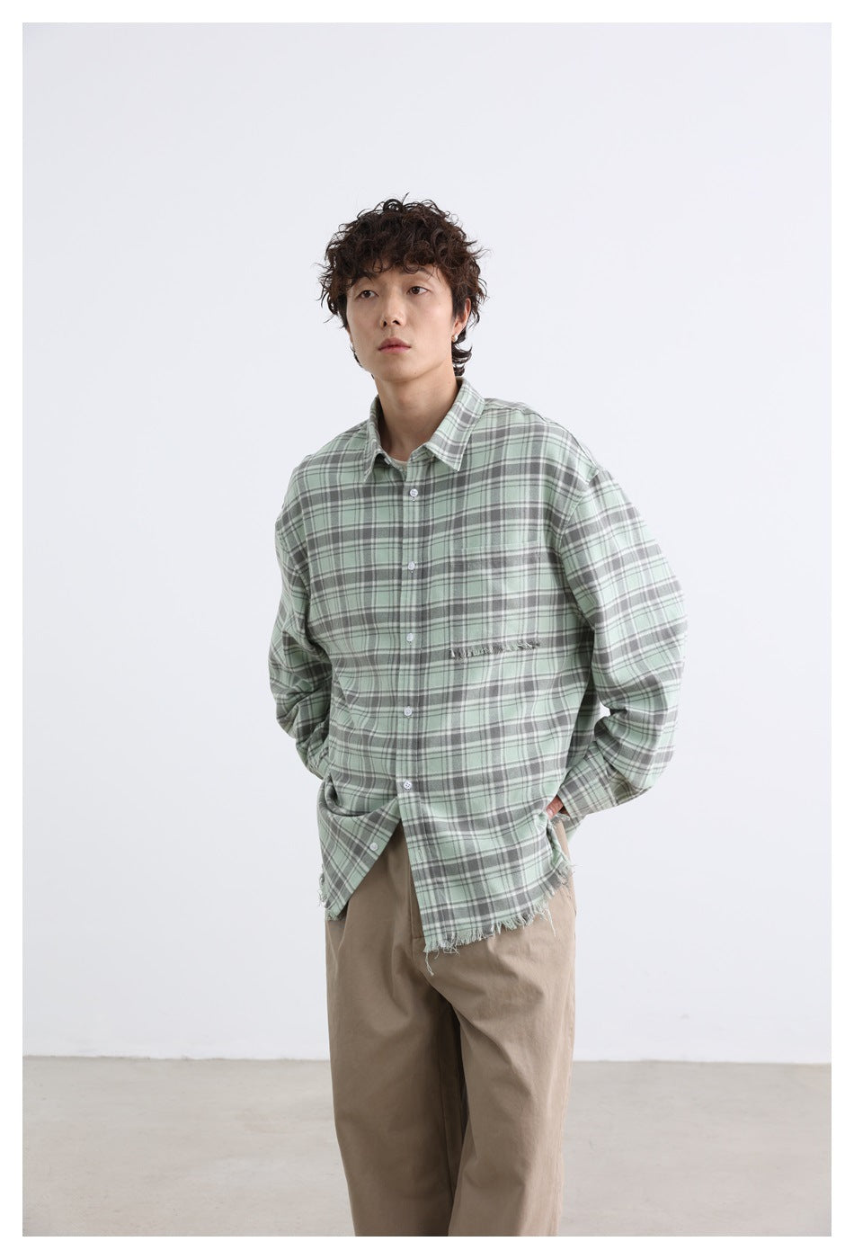 Model wearing a loose sage-green and gray plaid button-up shirt with a frayed chest pocket and hem.