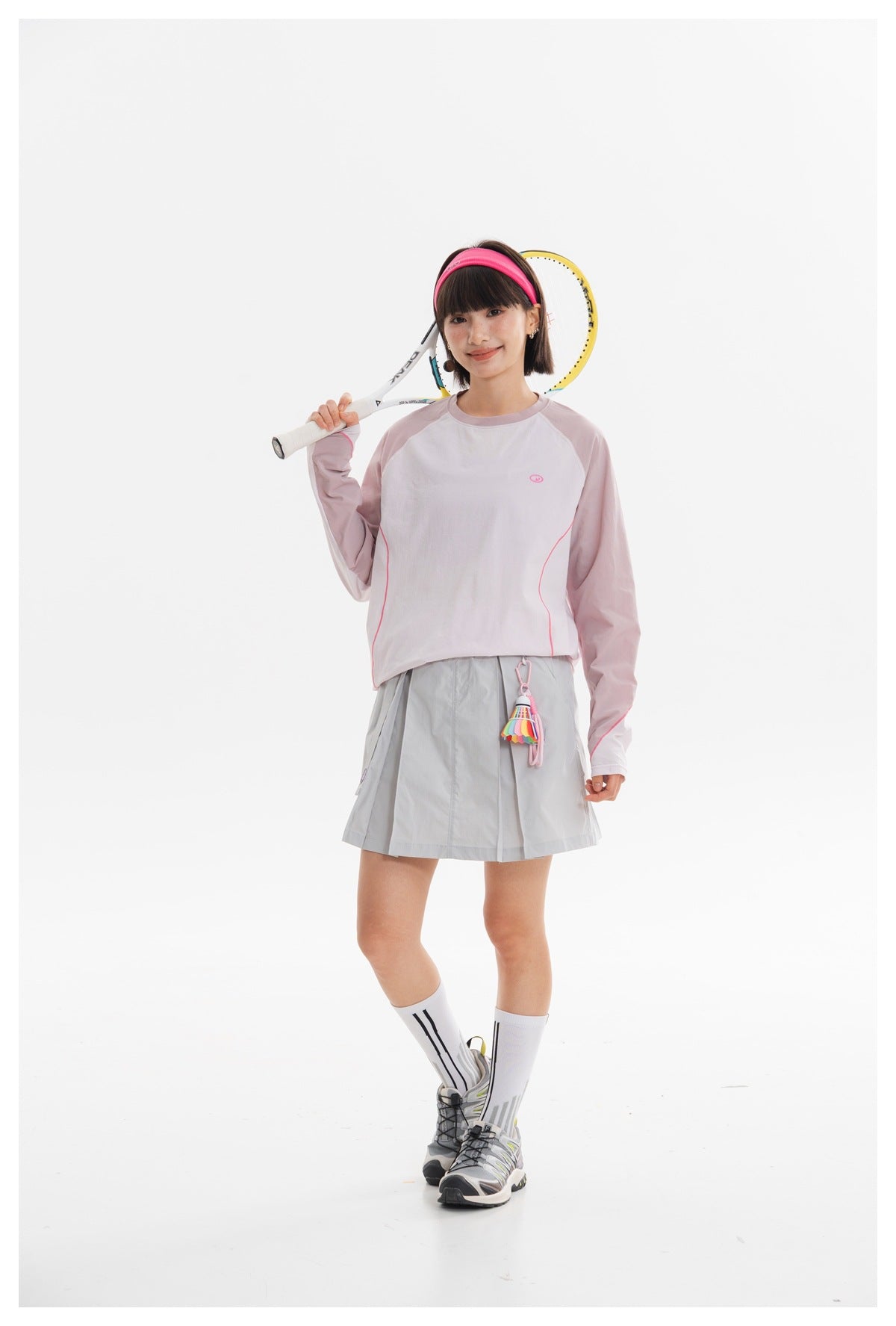 Model posing with a tennis racket, wearing a pale lavender long-sleeve top and a light gray pleated skirt.