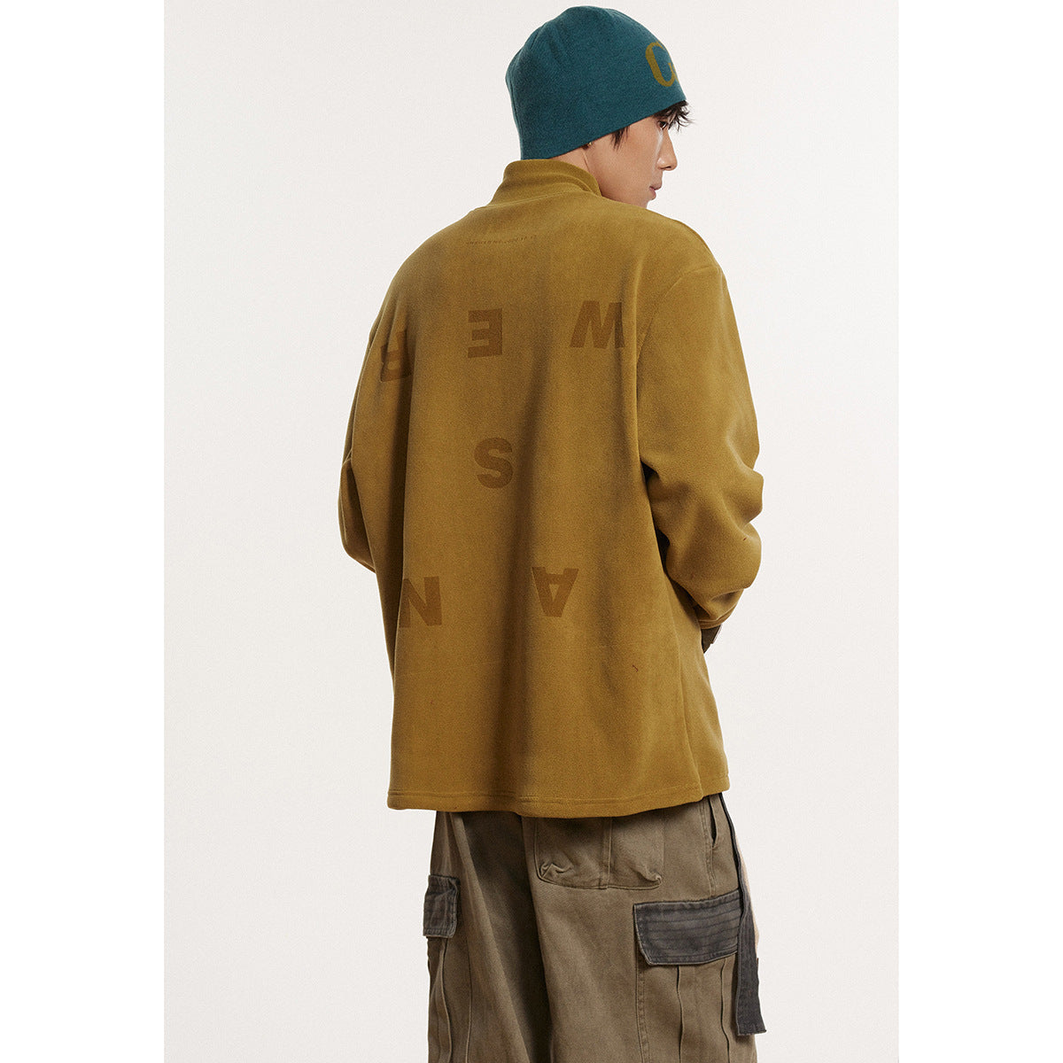 Model wearing a mustard-brown high-neck pullover with scattered back lettering and cargo pants, shown from behind.