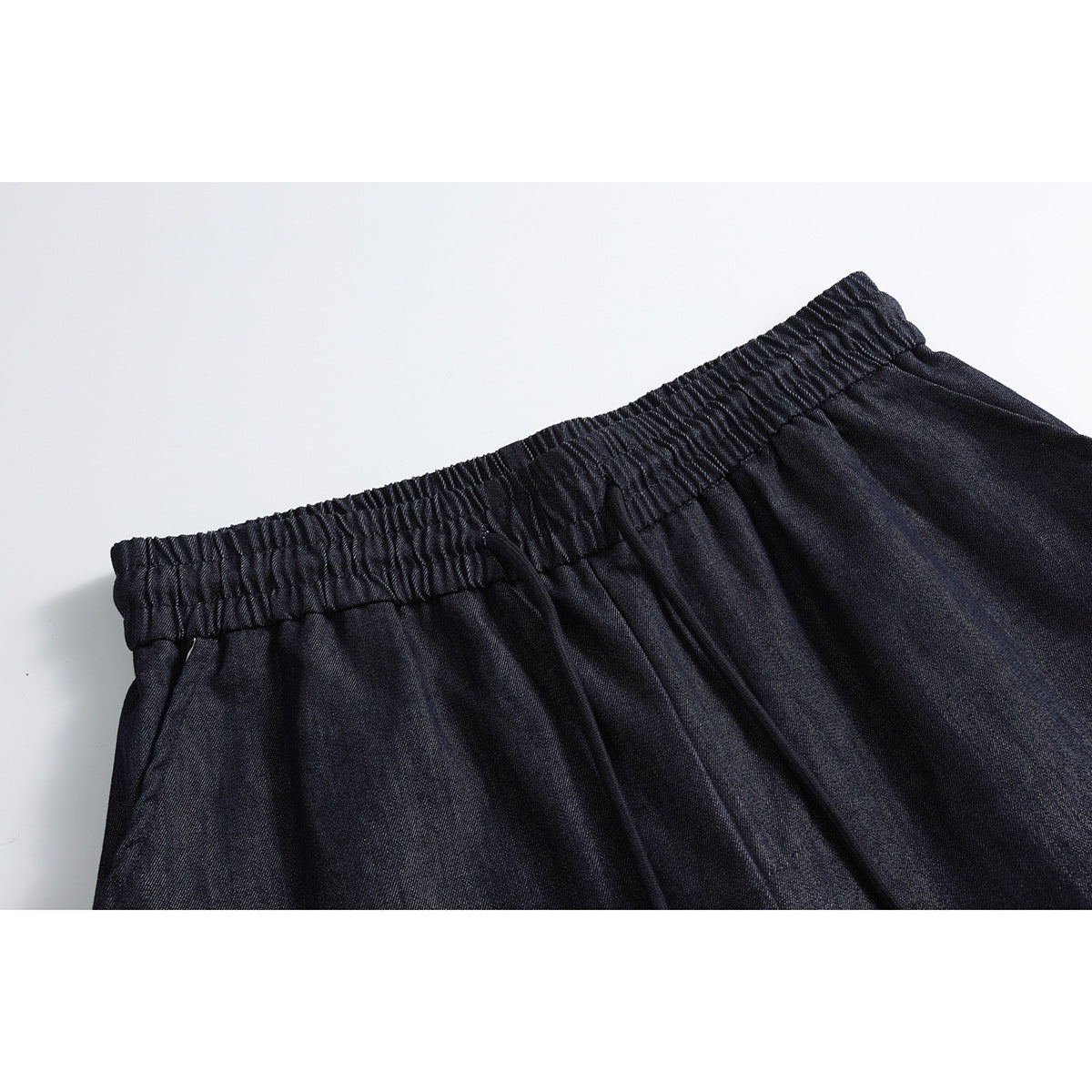 Close-up of dark indigo pants with a gathered elastic waistband, drawstring, and side pocket opening.