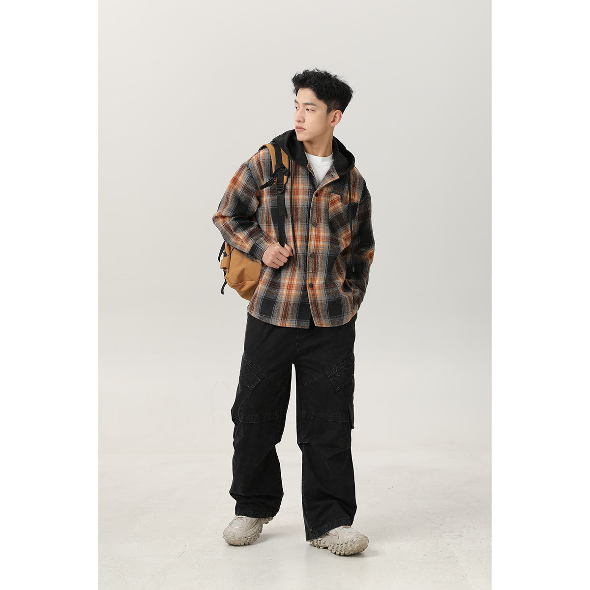 Man wearing a black, gray, and orange plaid hooded button-front overshirt with black cargo pants and a tan backpack.