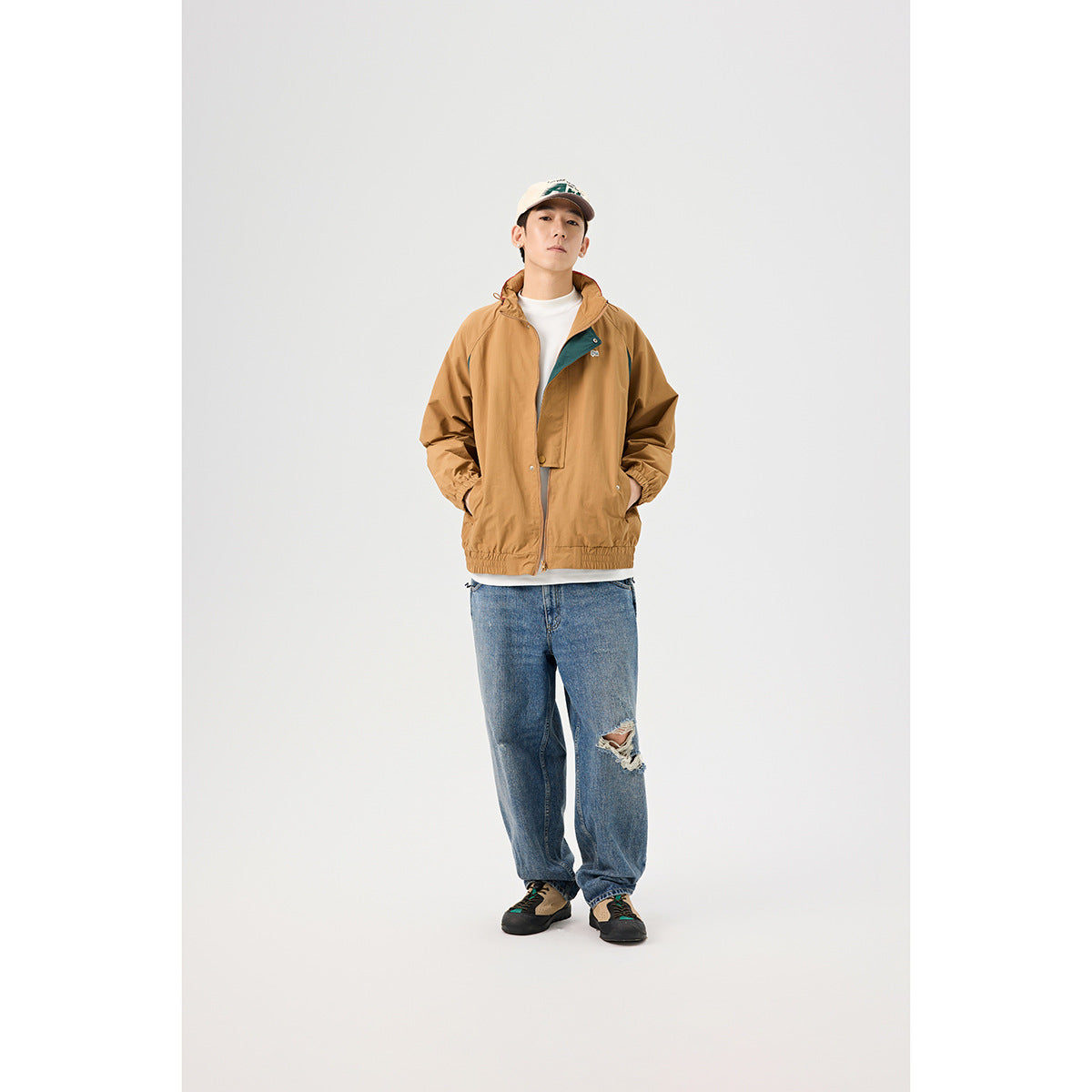 Front view of a model wearing a tan hooded jacket with dark teal accents and blue distressed jeans.