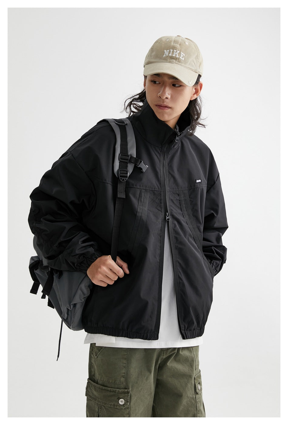 Model wearing a black zip-front jacket with a stand collar and elastic cuffs, styled with a backpack and khaki cap.