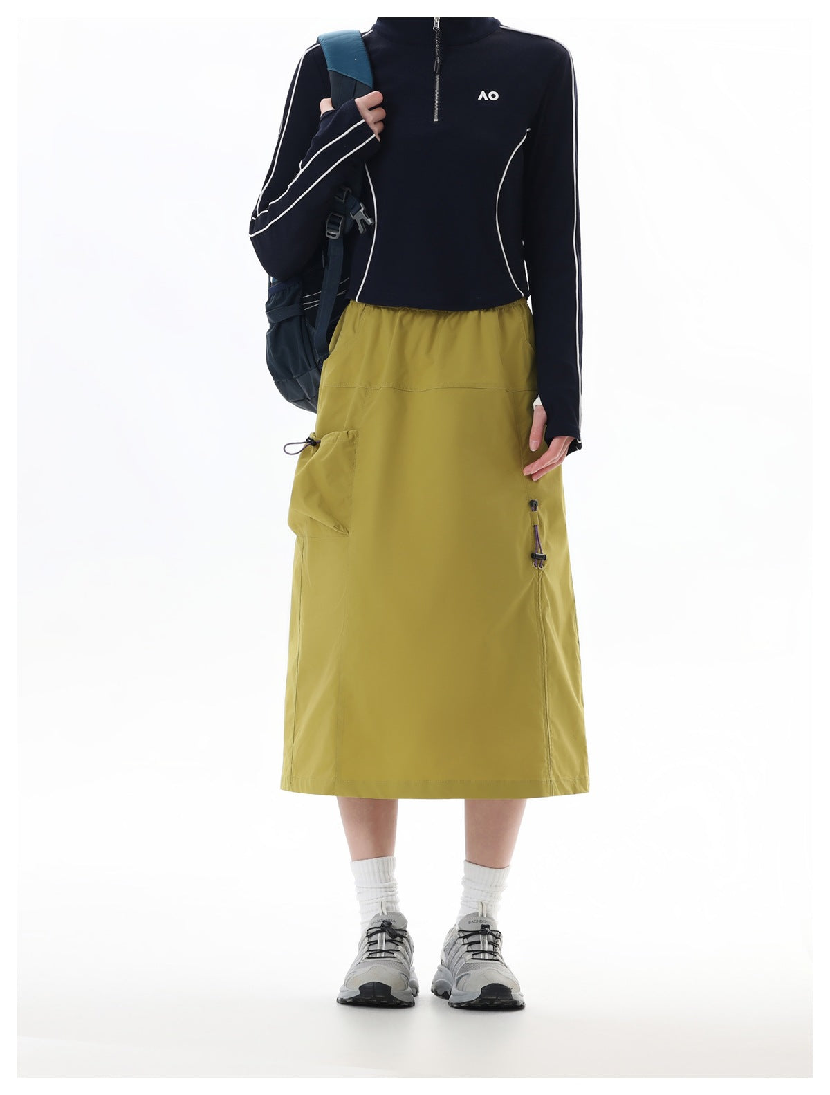 Model wearing a yellow midi skirt with side patch pocket, navy zip top, backpack and gray sneakers