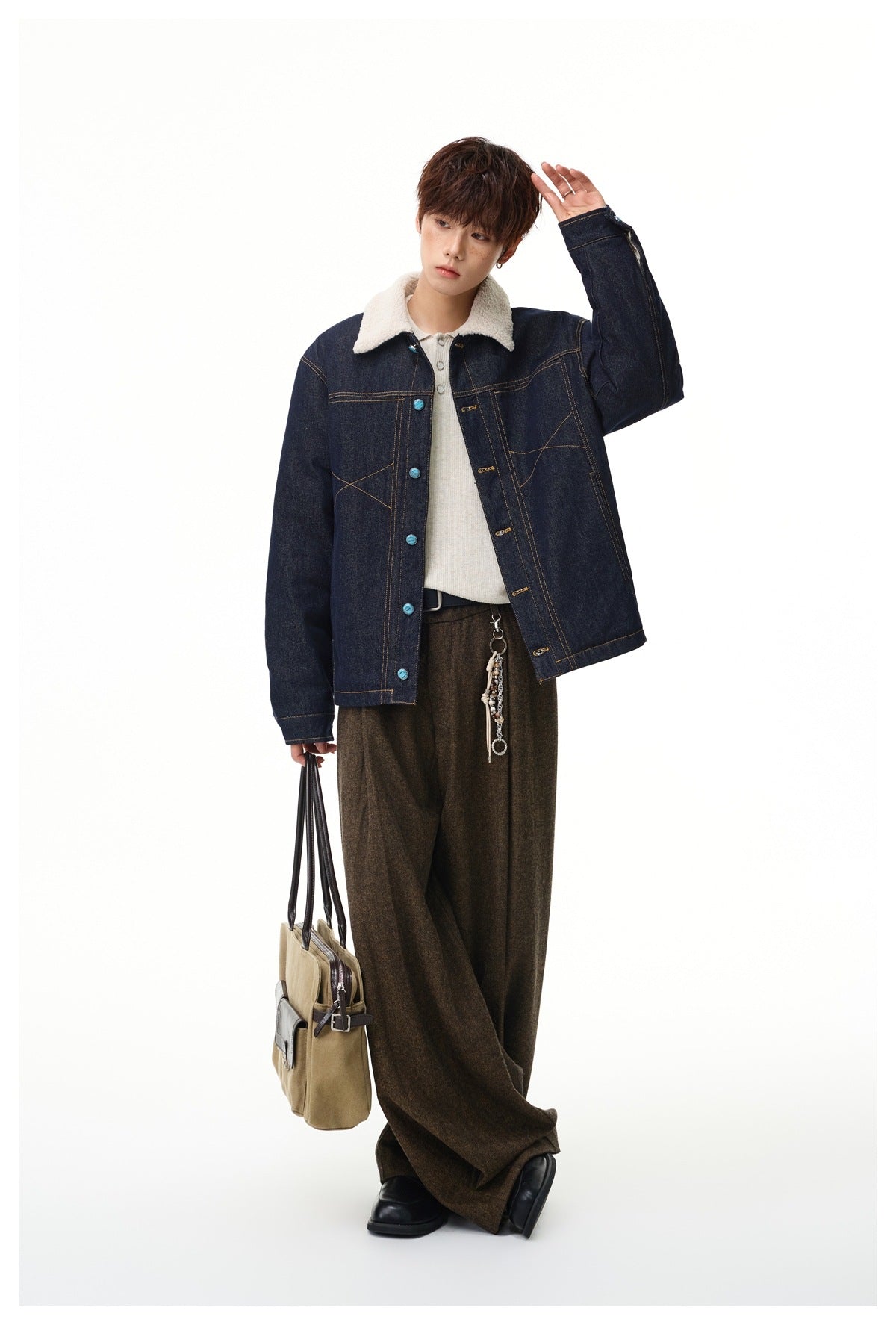 Model with one hand raised wearing denim jacket with cream fleece-like collar, brown wide-leg trousers and tan tote bag
