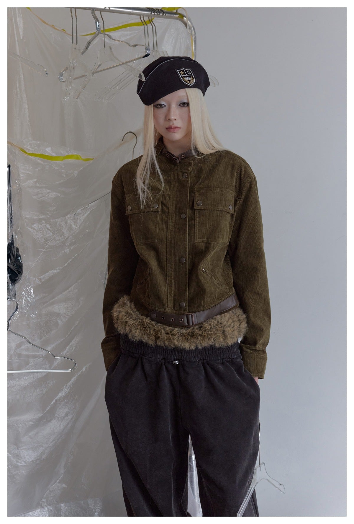 Close-up of a model wearing the olive corduroy jacket with buckled collar strap and flap pockets, with beret