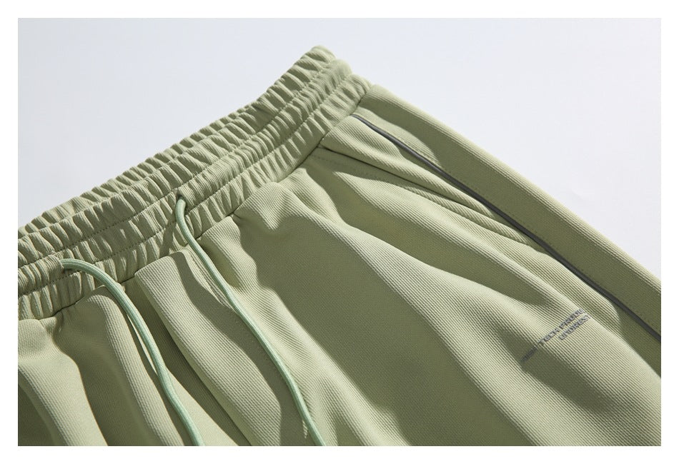 Close-up of pale sage pants showing the gathered waistband, drawstring, dark piping and small garment text.