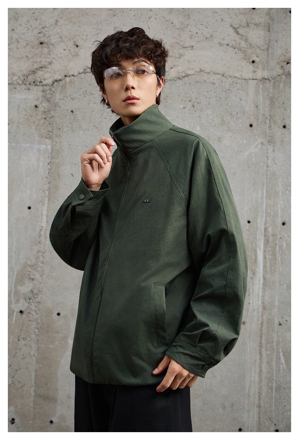 Man wearing an olive green stand-collar zip jacket with raglan sleeves and snap cuff tabs against a concrete wall