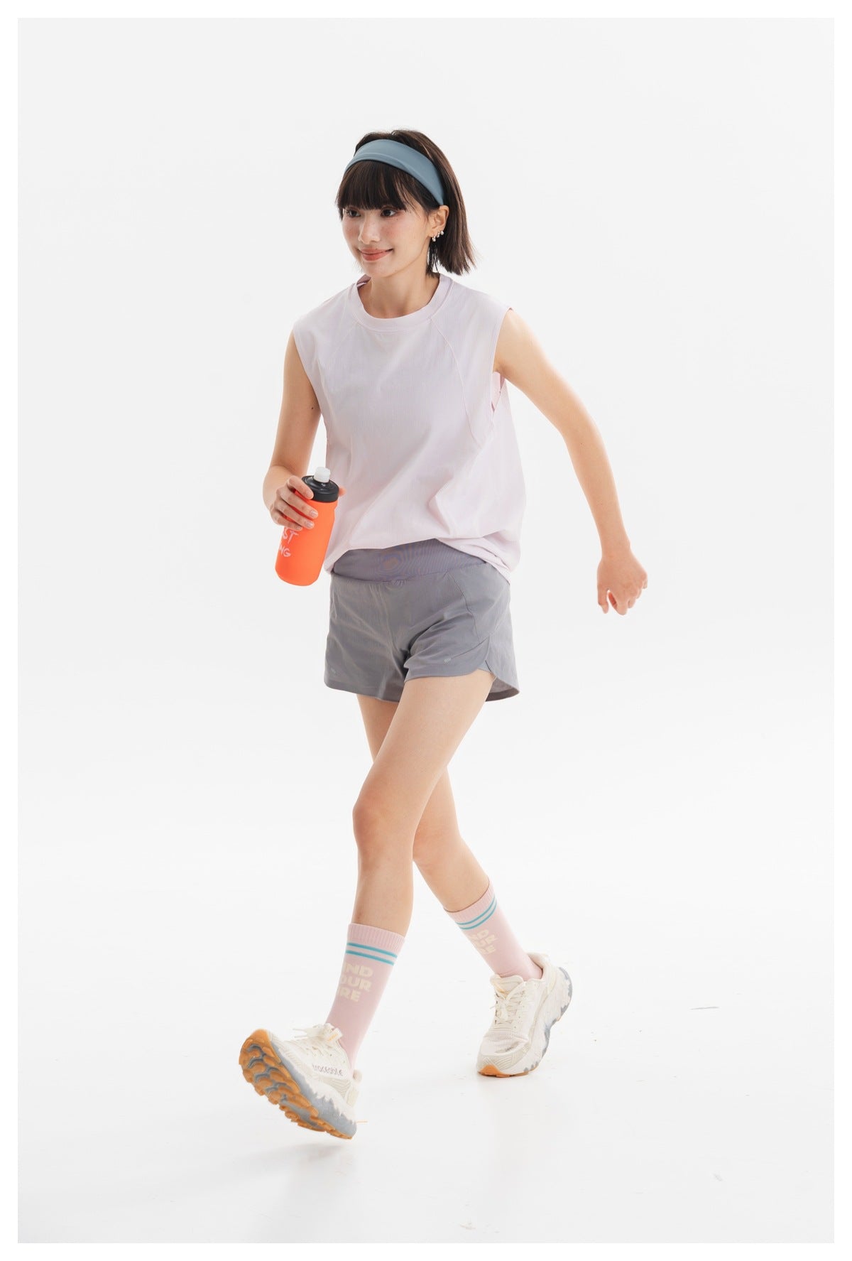 Model wearing a pale pink sleeveless athletic top and gray running shorts, shown walking in a bright studio.