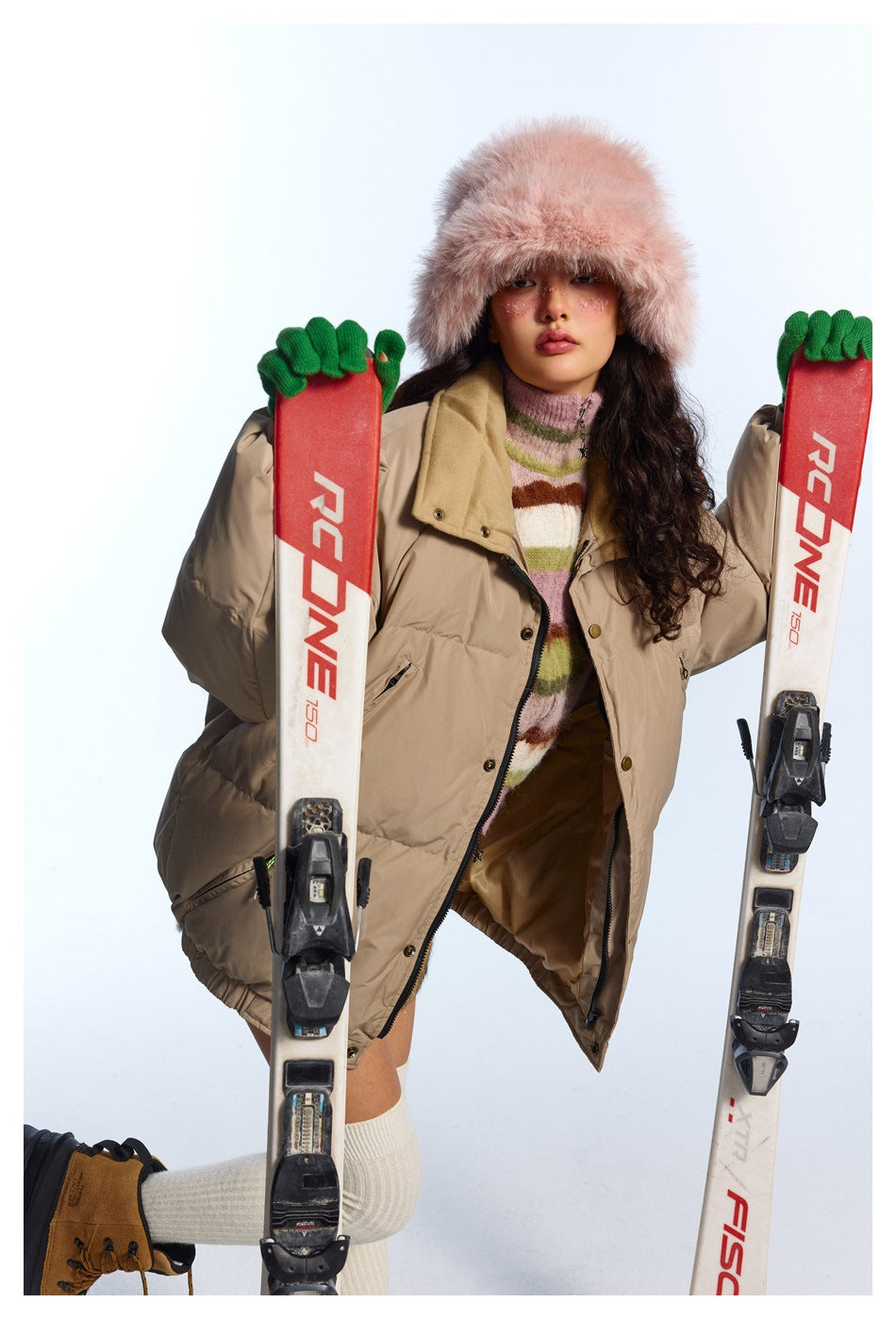 Model in an open beige puffer jacket and pink fur hat, holding two skis against a white background.