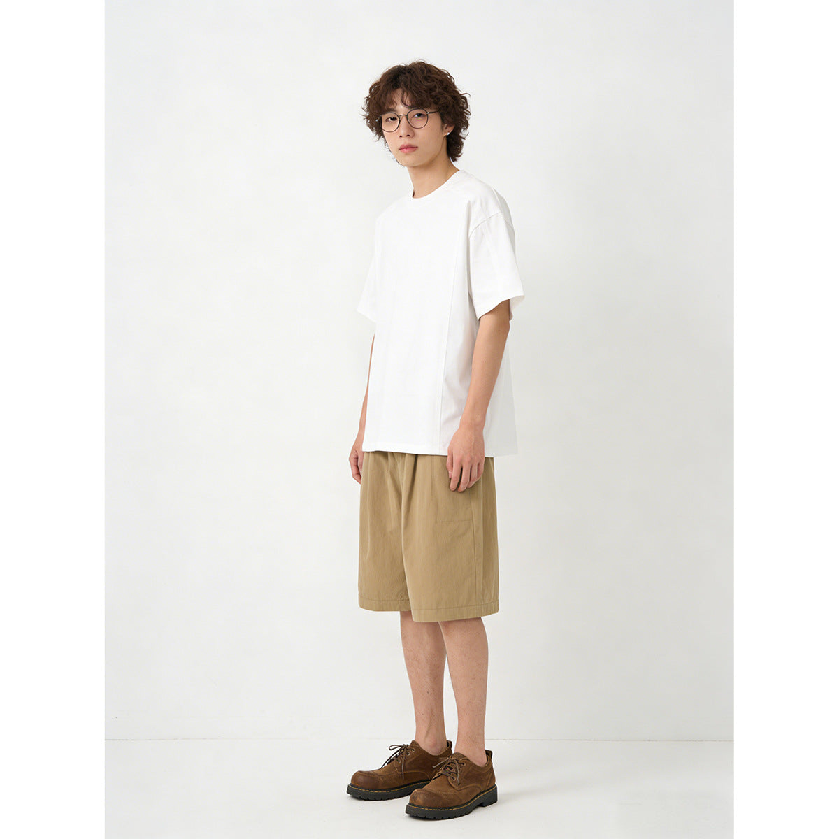 Model wearing a plain white short-sleeve T-shirt and tan shorts, shown in a three-quarter view with brown shoes.