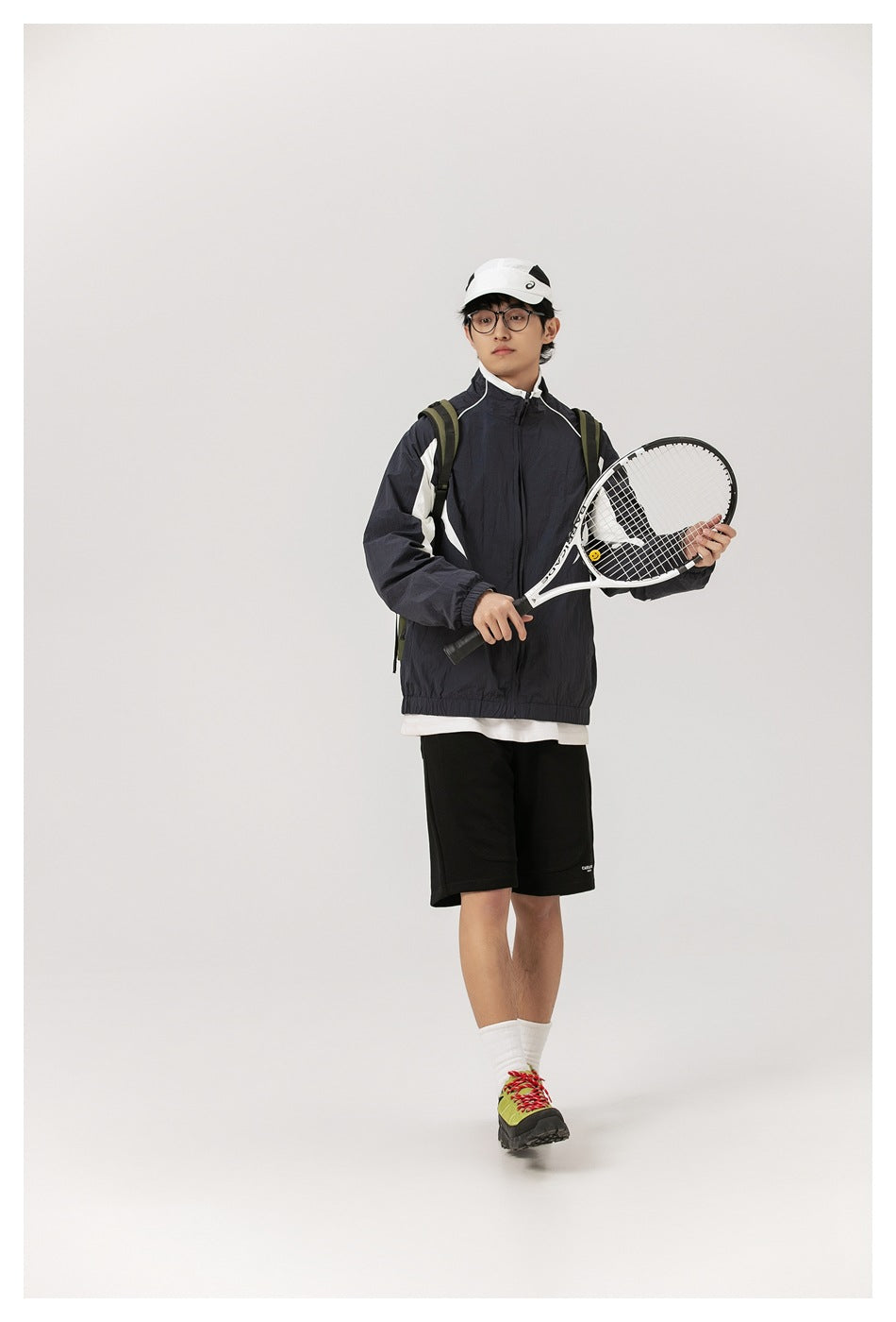 Full-length model wearing a zipped dark jacket with white panels and black shorts, holding a tennis racket.