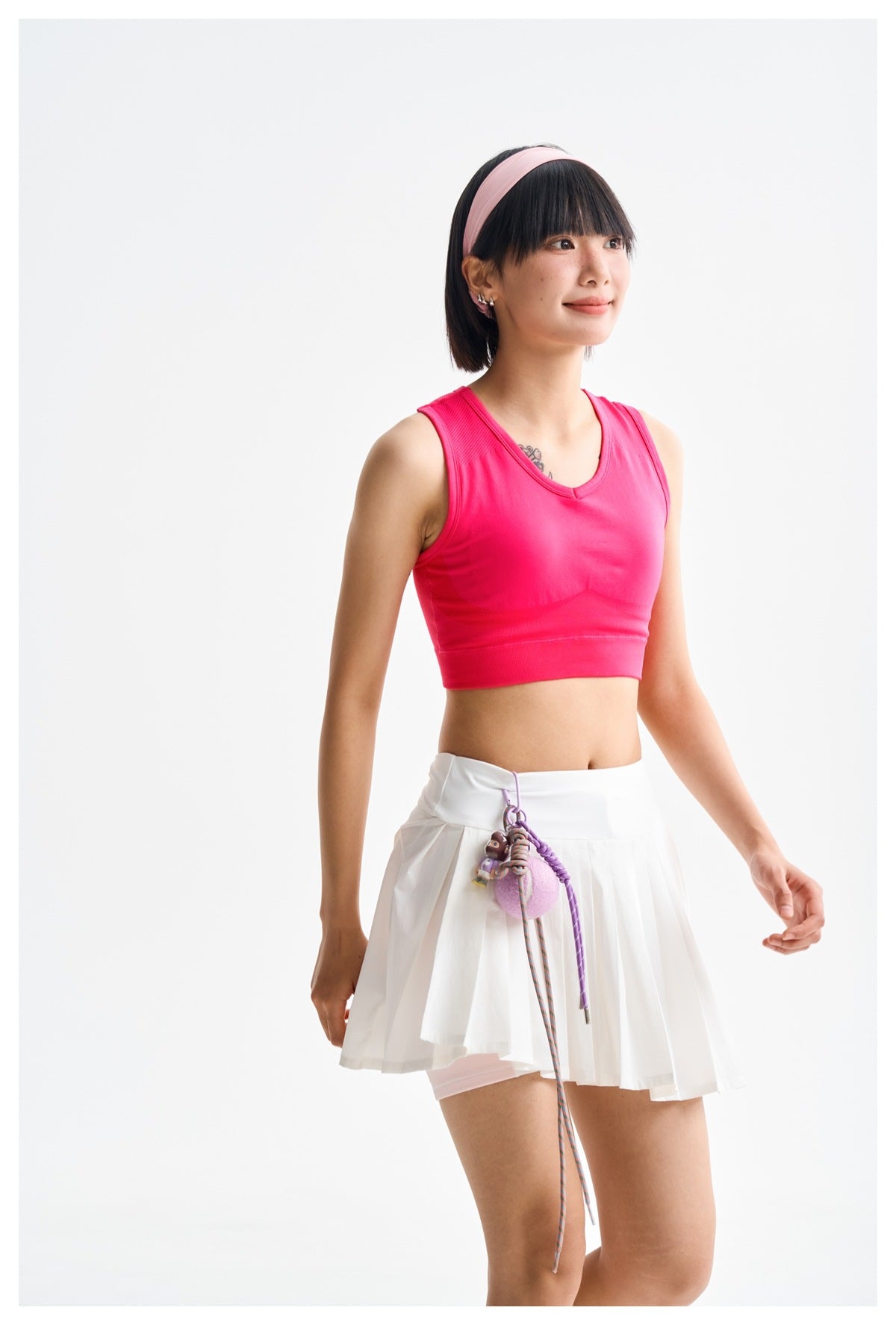 Model in a hot-pink sleeveless crop top and white pleated tennis skirt, shown at a three-quarter angle.
