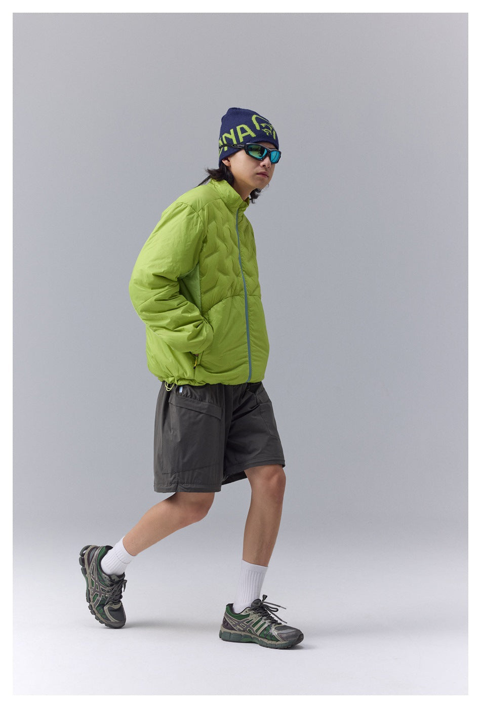 Model wearing a lime green quilted zip jacket with gray shorts, beanie, sunglasses and sneakers, walking with hand in pocket