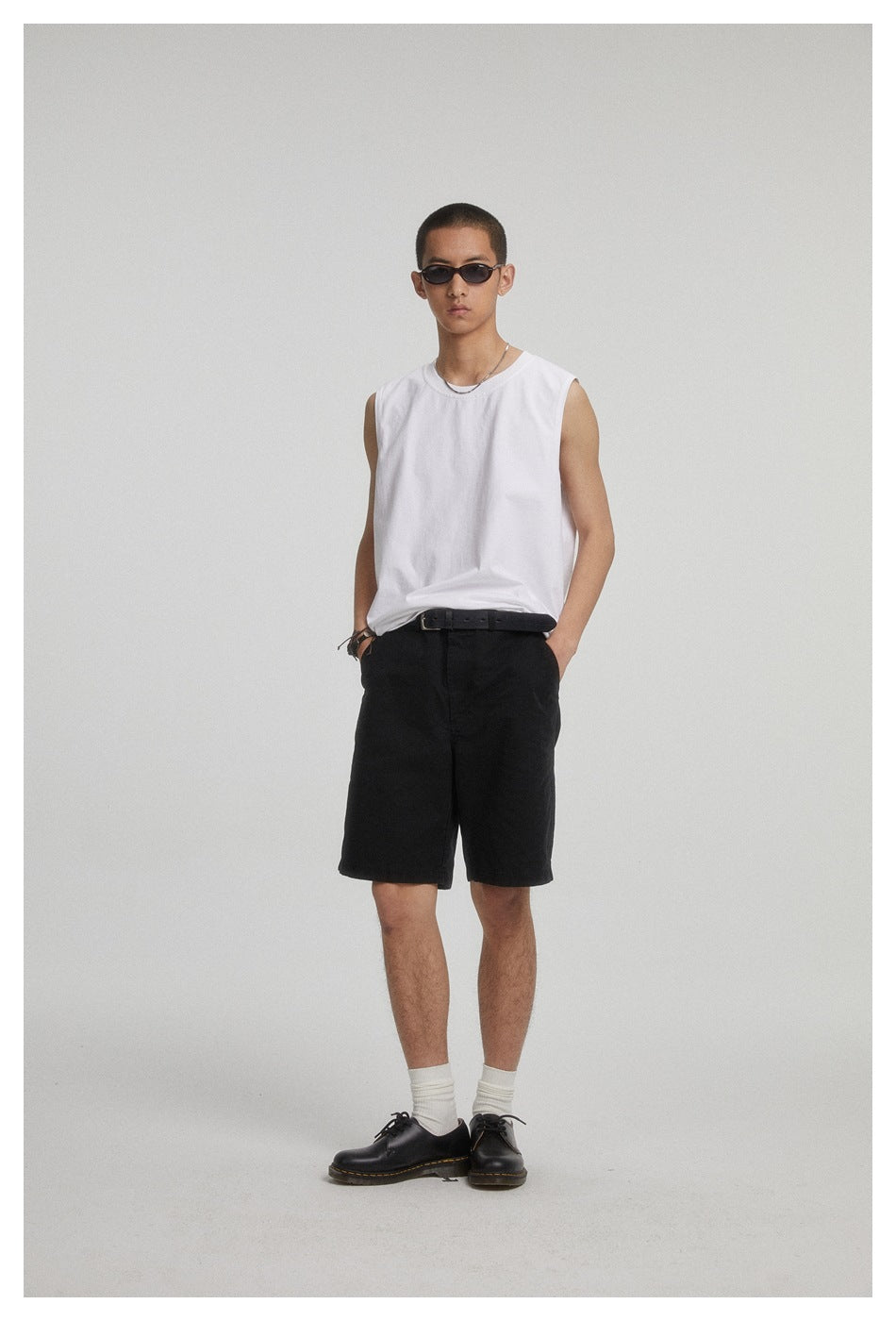 Full-length model wearing a plain white sleeveless crew-neck shirt, black shorts, white socks, and black shoes.