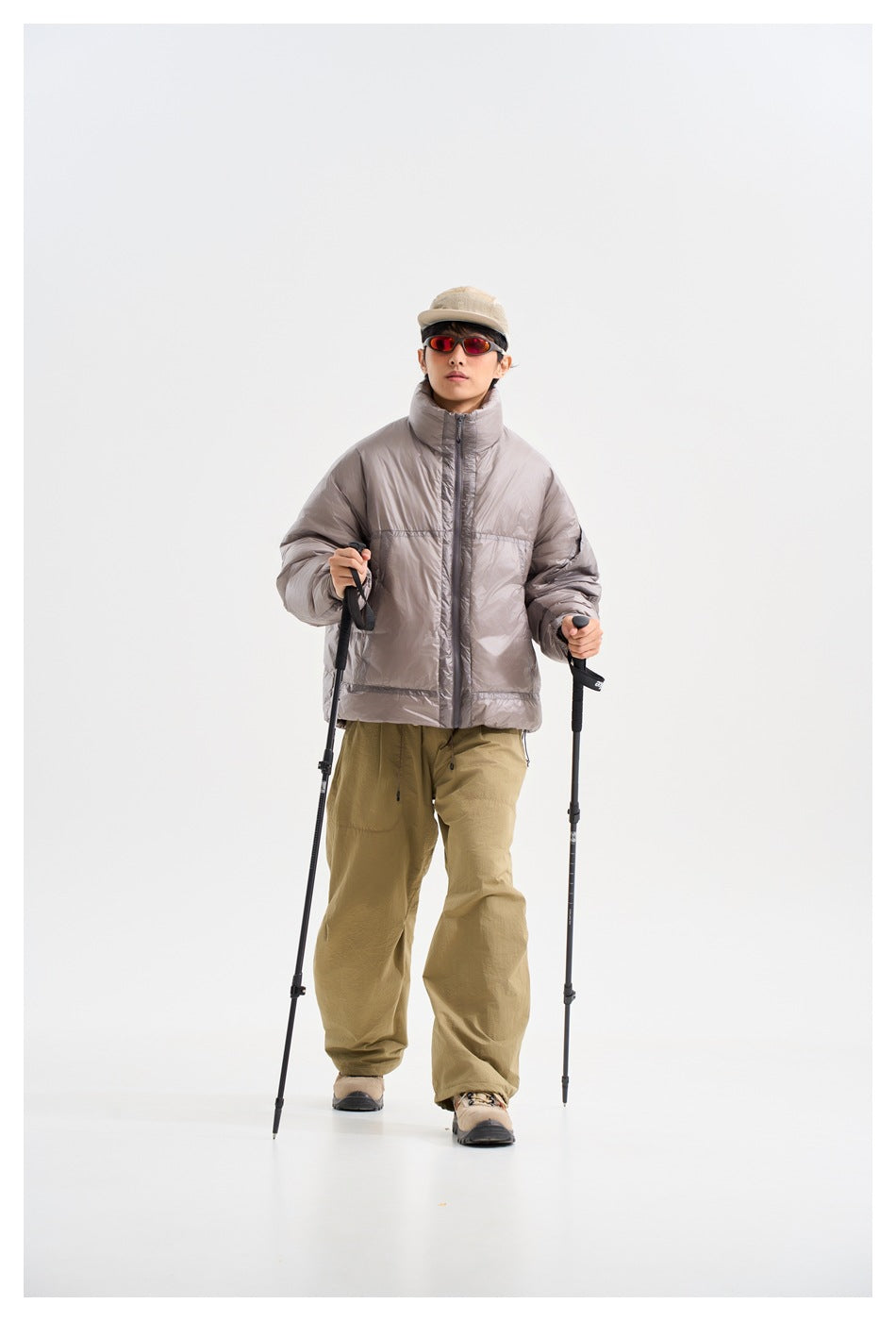 Model wearing a light gray padded jacket and khaki trousers, holding trekking poles and facing forward.