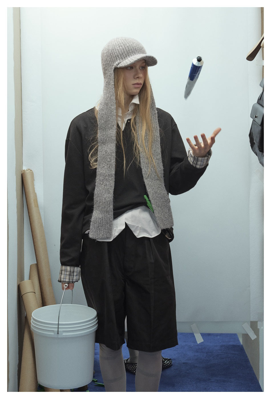 Front three-quarter view of a model in a black top and knee-length shorts, white shirt, and long gray scarf and cap.