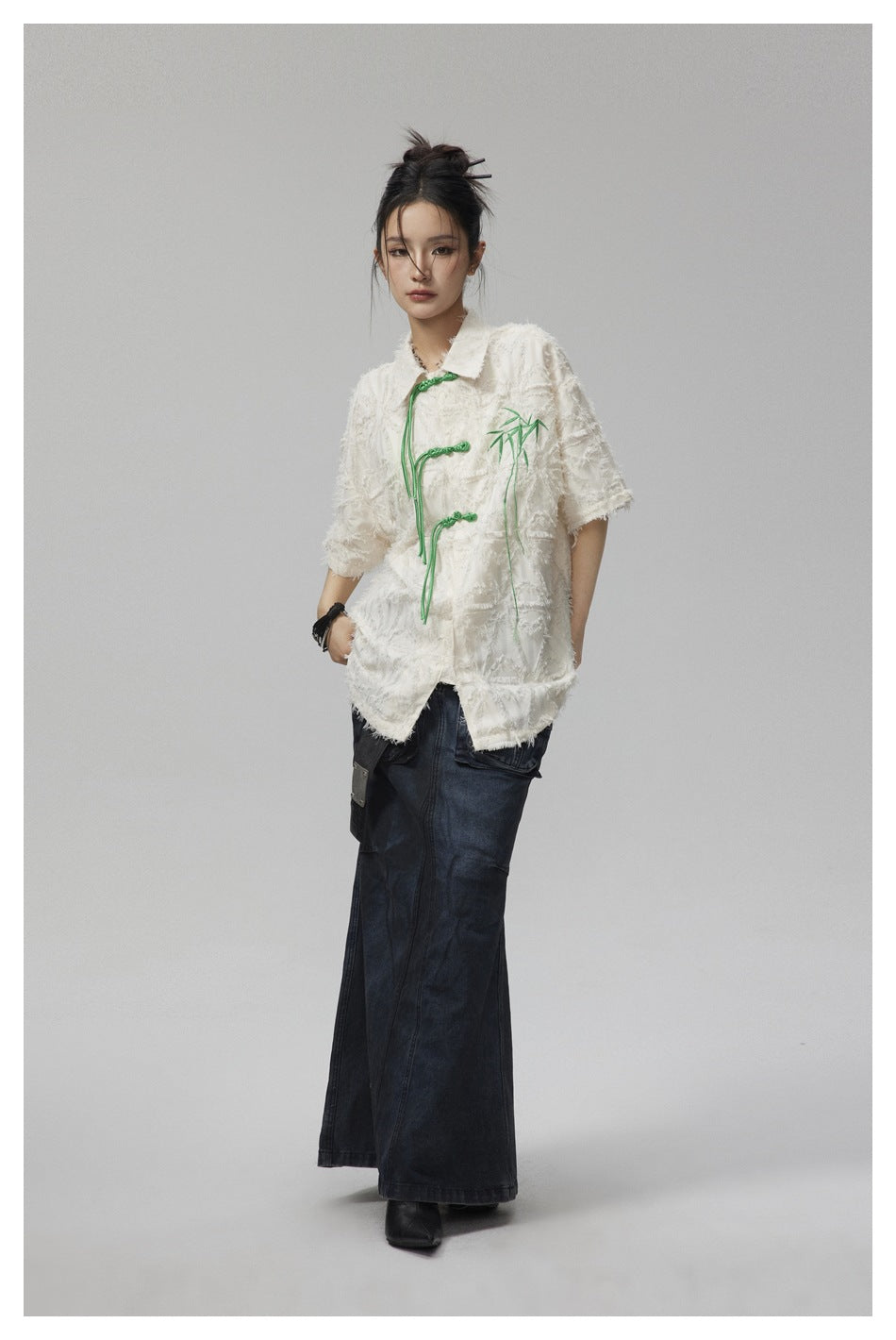 Front view of a model in an ivory textured jacket with green frog closures and a dark denim maxi skirt.