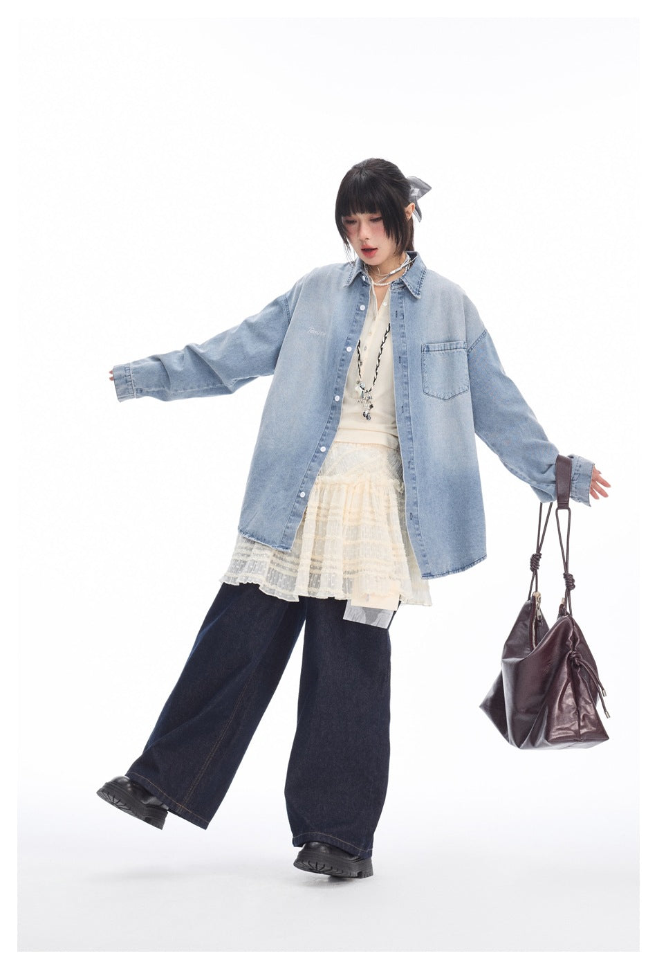 Full-length model wears a light blue oversized denim shirt, cream lace layers and dark wide-leg jeans against a white