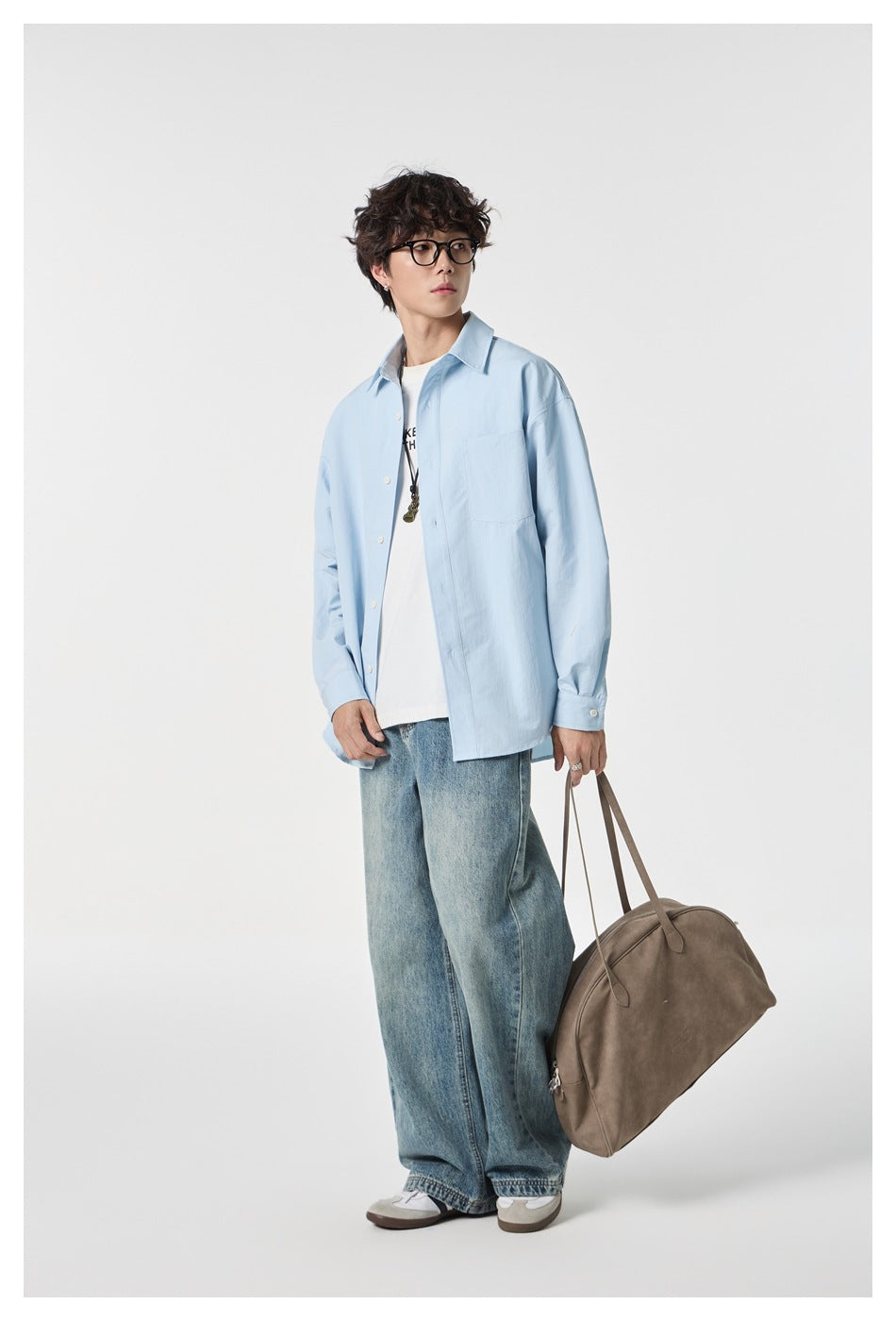 Full-length view of a model in an oversized pale blue button-up shirt, white tee, and loose jeans, carrying a taupe bag.
