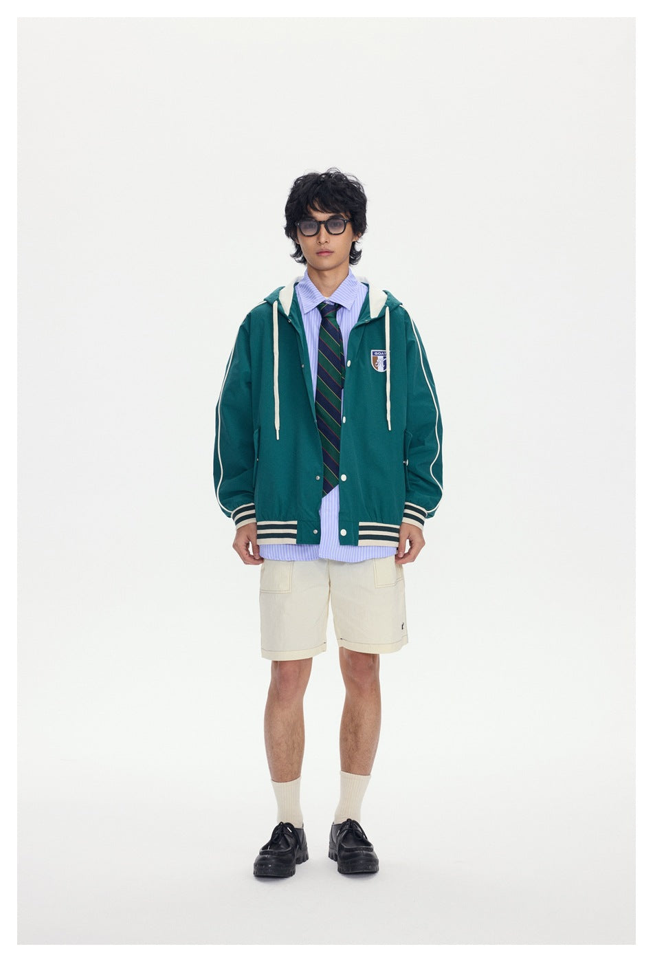 Model wearing a dark green hooded snap-front jacket over a striped shirt and tie with cream shorts, hood down