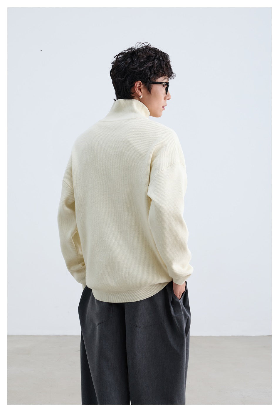Model seen from behind wearing an ivory high-neck pullover with dropped shoulders and dark trousers.