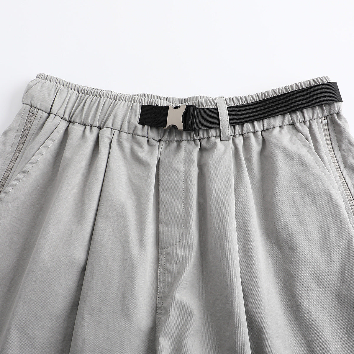 Close-up of light gray shorts with an elastic waist, black webbing belt and buckle, belt loop, and front pockets.