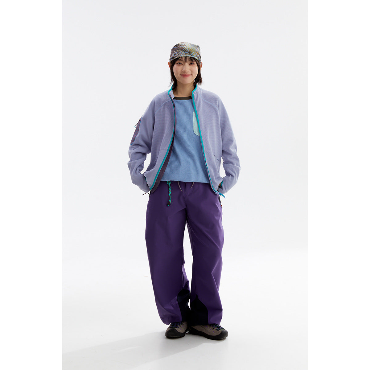 Model wearing a lavender zip jacket with turquoise trim, blue top, loose purple pants and a patterned cap.