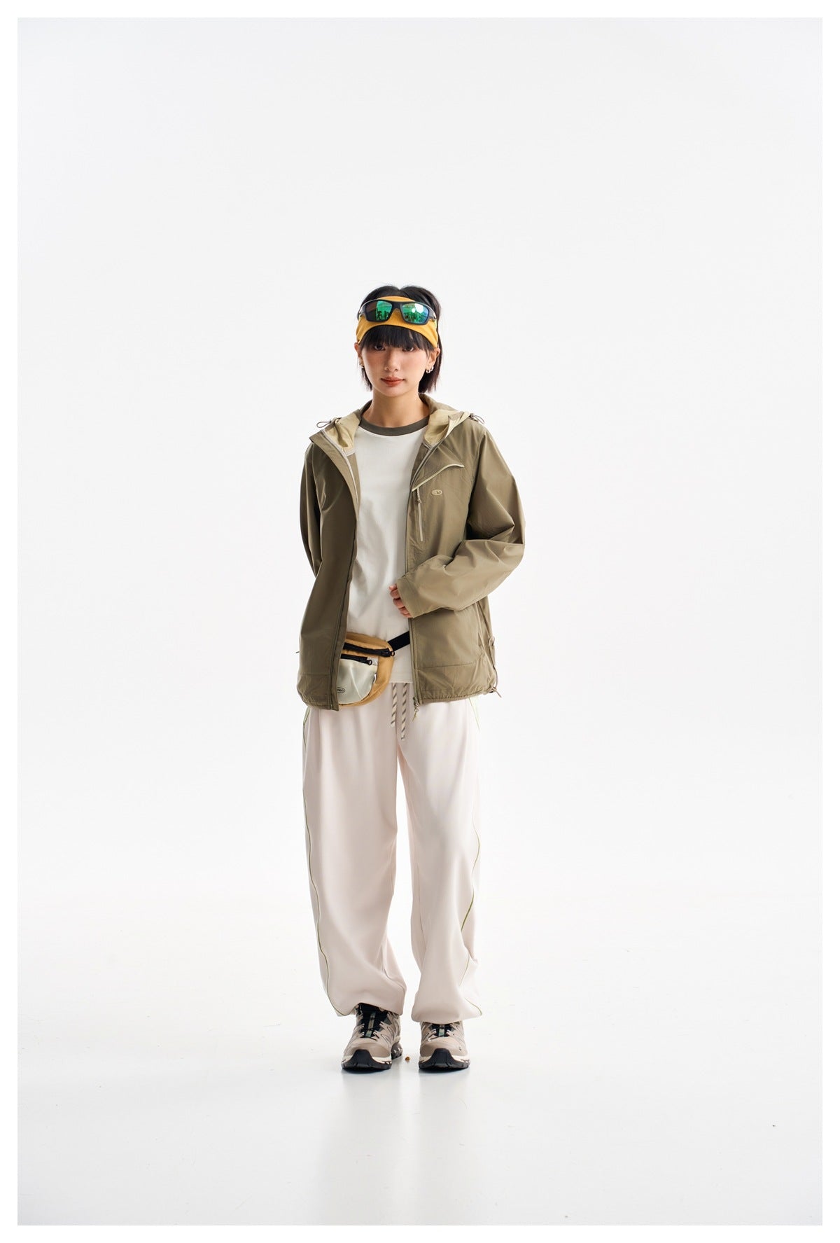 Model wearing an olive hooded jacket open over a white shirt, with loose light pants, shown from the front.