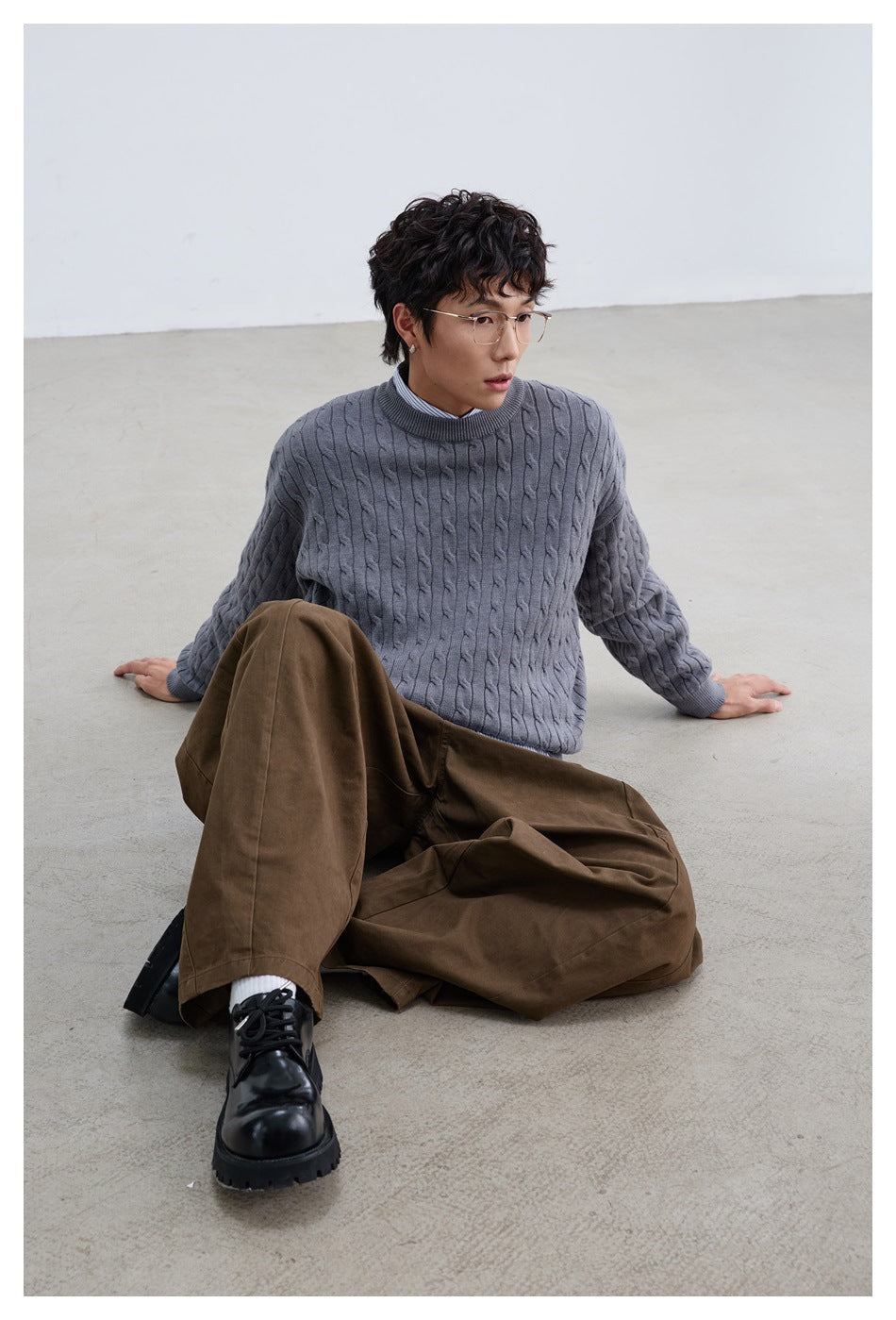 Model seated on floor wearing gray cable-knit sweater over a striped shirt with brown wide-leg pants and black shoes