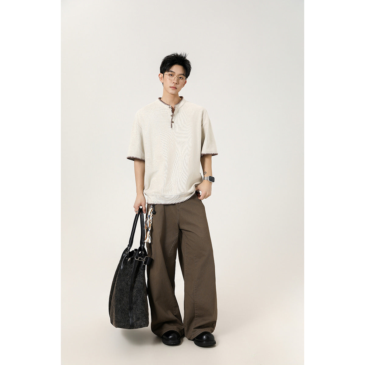 Model wearing an off-white short-sleeve henley with dark contrast stitching, brown wide-leg trousers and black shoes.