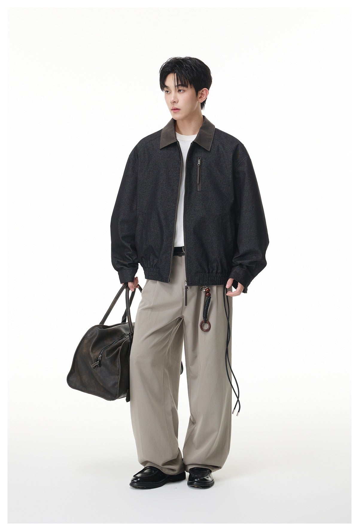 Model wearing a black-gray zip jacket with brown collar, white tee and wide taupe trousers, holding a leather bag