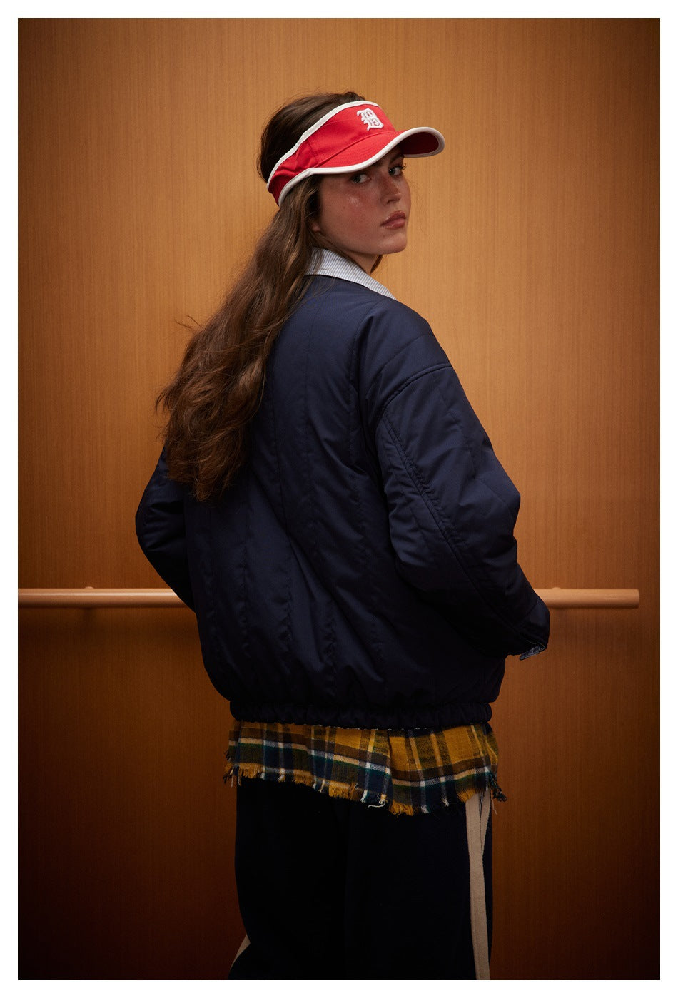 Model in a navy oversized jacket, shown from behind, with a red visor and a plaid skirt.