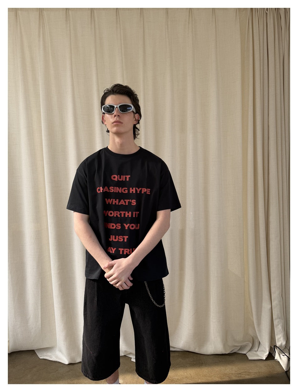 Model wearing a black T-shirt with red text, black shorts and sunglasses, shown against beige curtains.