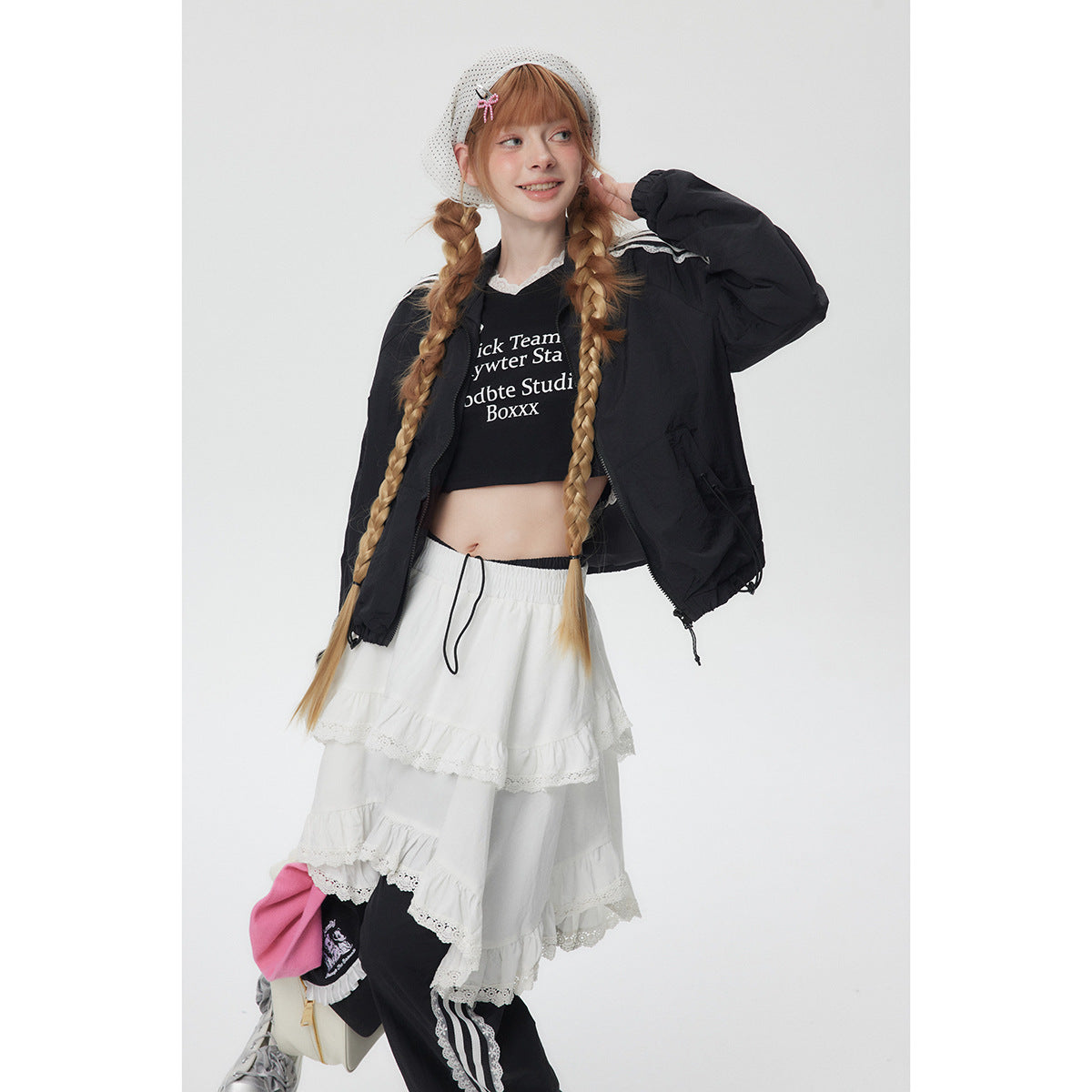 Model wearing a black zip jacket over a printed crop top, white tiered lace skirt and black track pants.