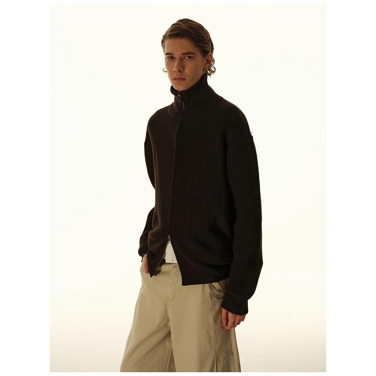 Model wearing a dark ribbed zip-up cardigan with a high collar and loose beige trousers.