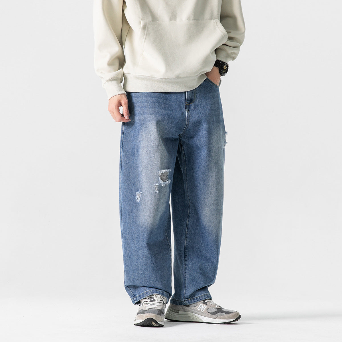 Front view of a model wearing loose blue jeans with distressed patches, a cream sweatshirt, and gray sneakers.