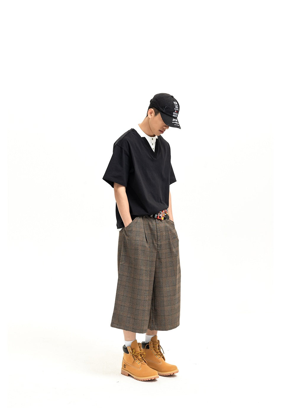 Model in black layered-look V-neck tee with white collar, cap, plaid wide-leg shorts and boots, full-length side view