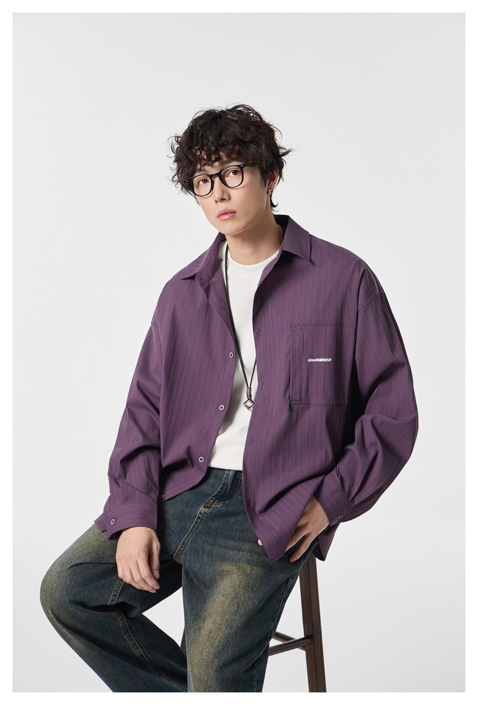 Seated model in a muted purple button-front shirt with a chest pocket, white tee, and wide-leg faded jeans.
