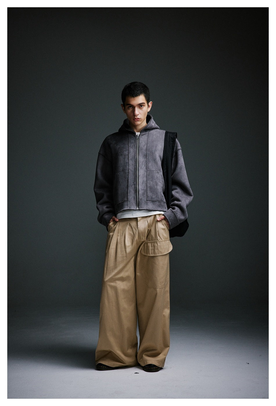 Model standing in khaki wide-leg pleated pants with a flap pocket, gray zip hoodie and black shoulder bag, hands in pockets