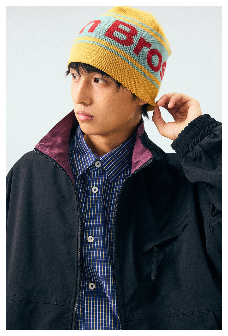 Close view of a model in a black jacket with plum collar lining and a blue checked shirt, wearing a yellow lettered beanie.