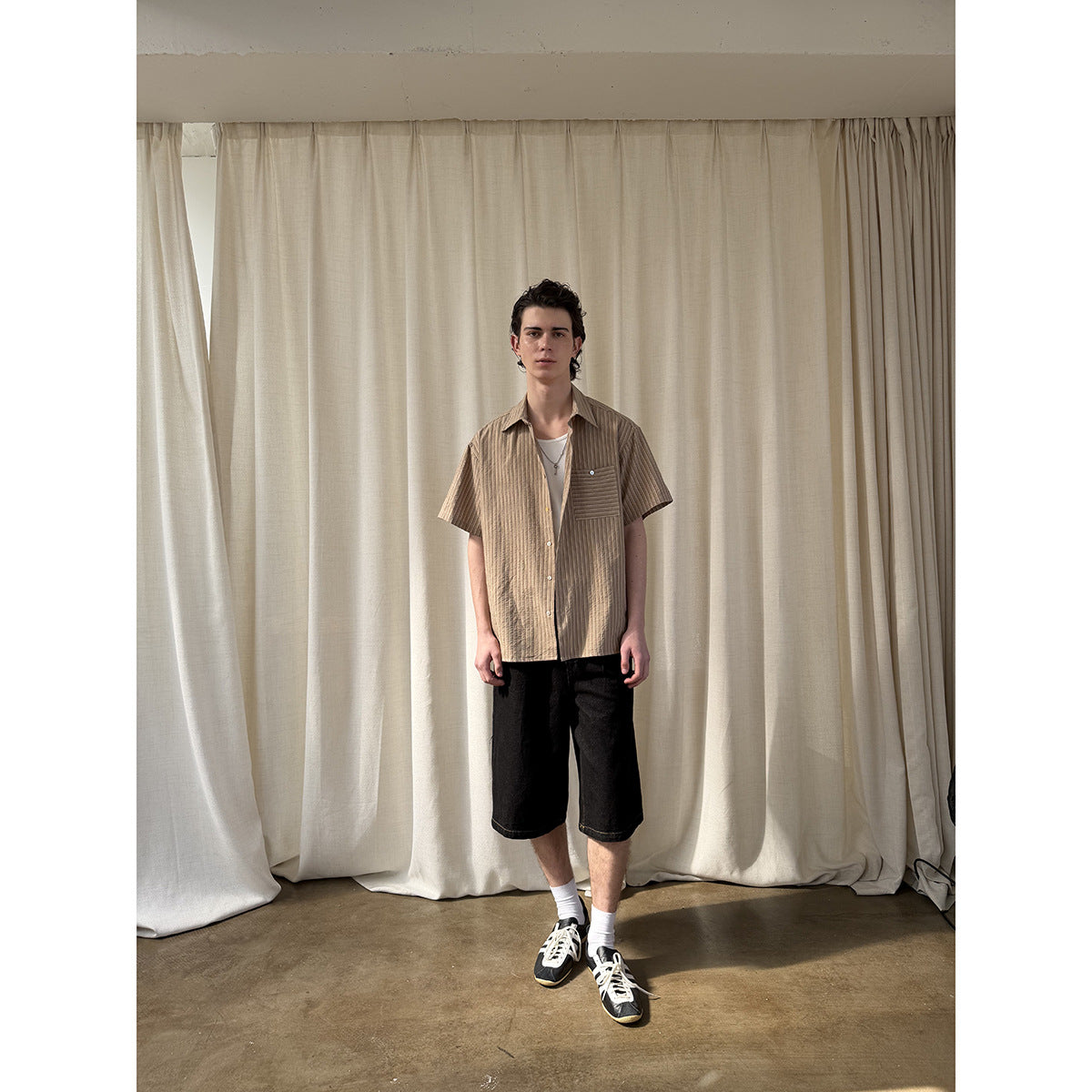 Full-length model wearing a tan vertically striped short-sleeve button-up shirt with chest pocket and black shorts.