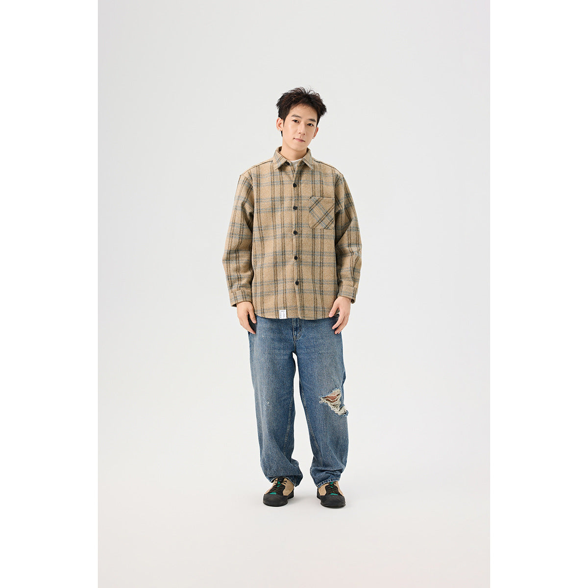 A man models a tan plaid button-front overshirt with a chest pocket, blue jeans, and sneakers.
