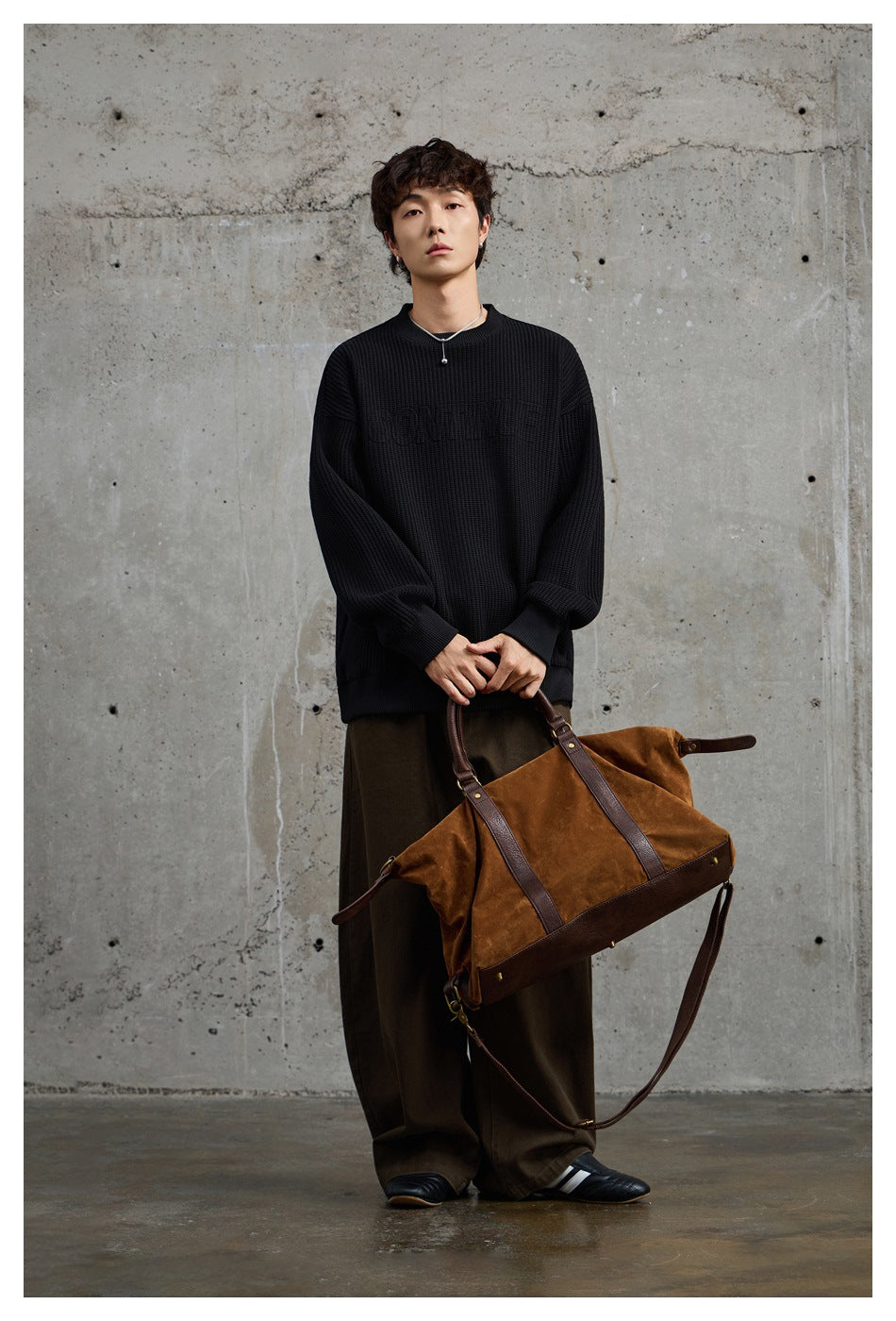 Front view of a model in an oversized black textured crewneck sweatshirt, holding a brown leather-trimmed bag.