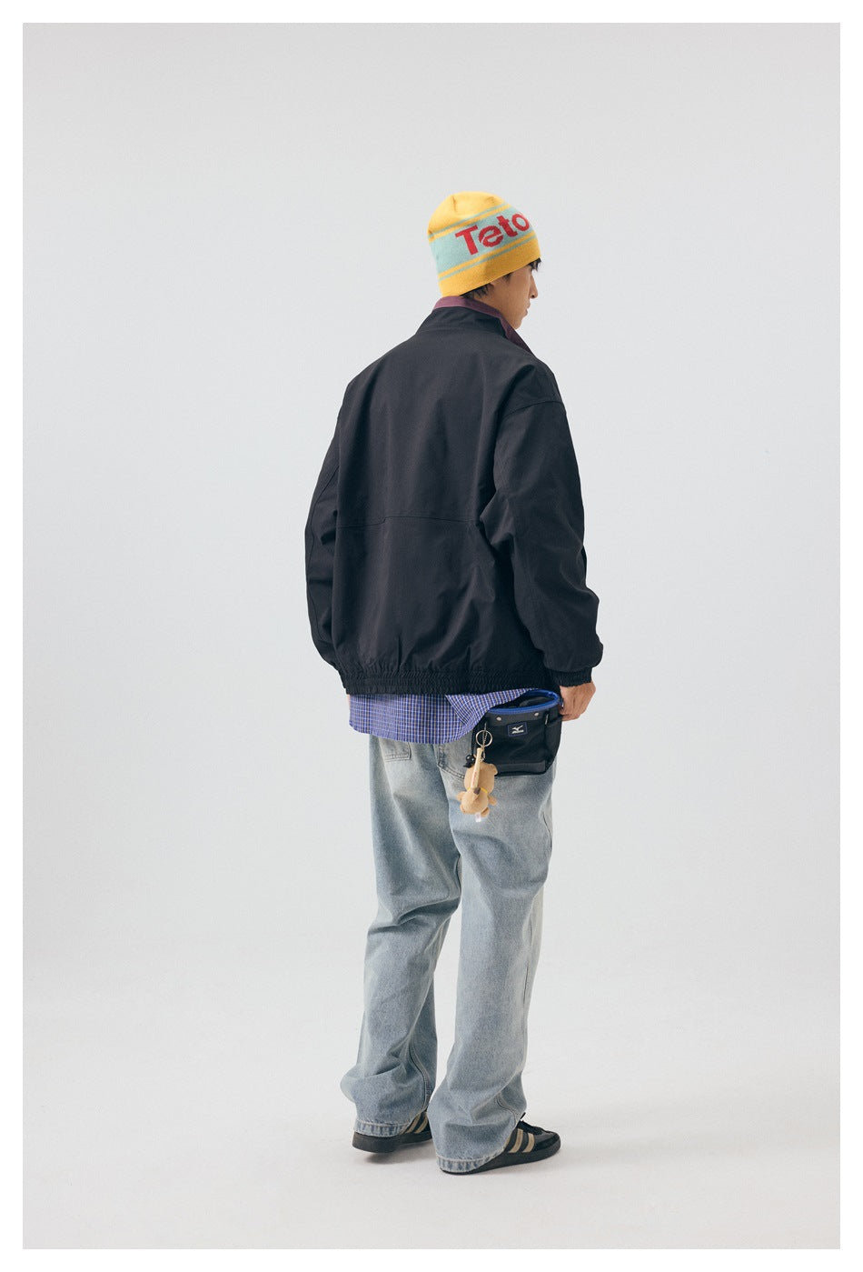 Back view of model wearing black jacket with elastic hem over a plaid shirt, light wash jeans and a small bag