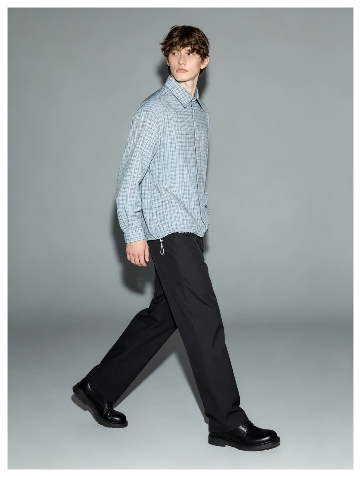 Model walking in a blue plaid button-up shirt with drawcord hem, black trousers and black lace-up boots