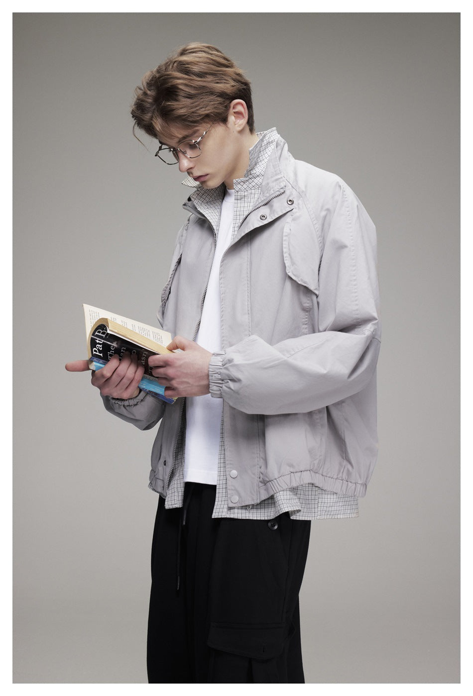 Model wearing a light gray zip-front jacket over a plaid shirt, holding an open book.