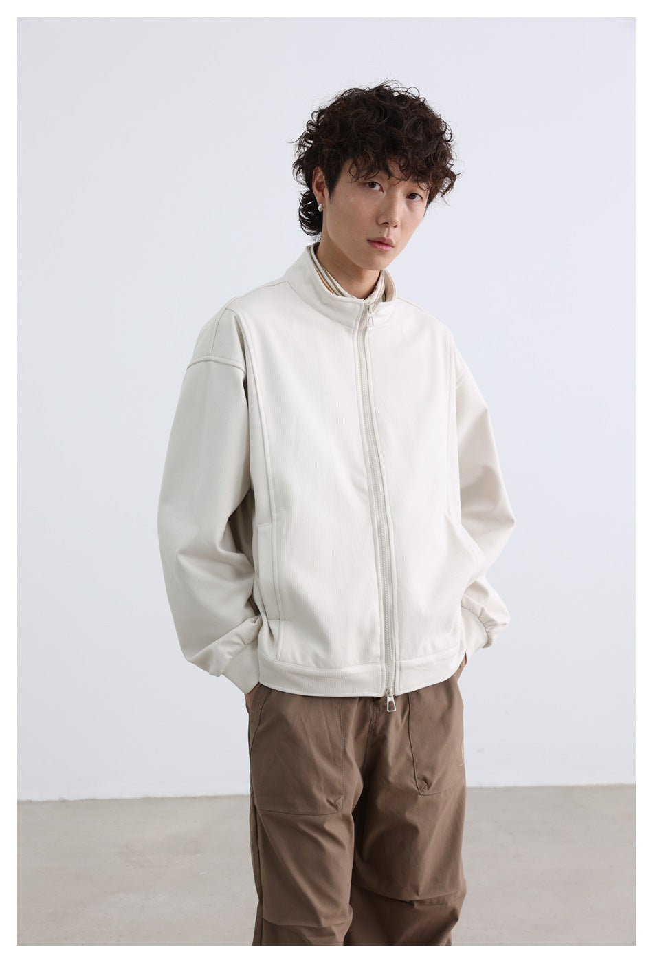 Front view of a model wearing an off-white zip-front jacket with a stand collar and taupe trousers.