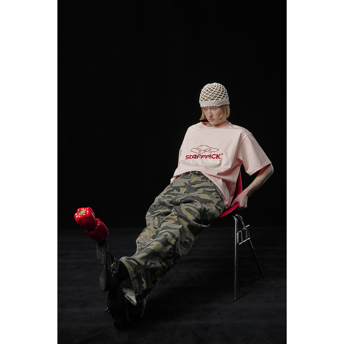 Seated model in a pale pink graphic T-shirt and loose camouflage trousers, wearing a cream knit cap.
