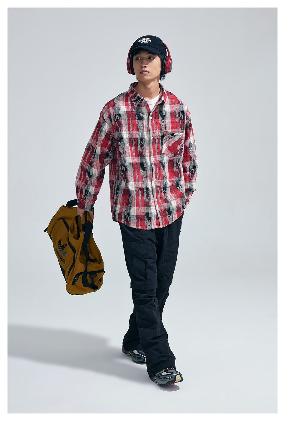 Model walking in red plaid button-up shirt over a white tee with black cargo pants, carrying a mustard bag